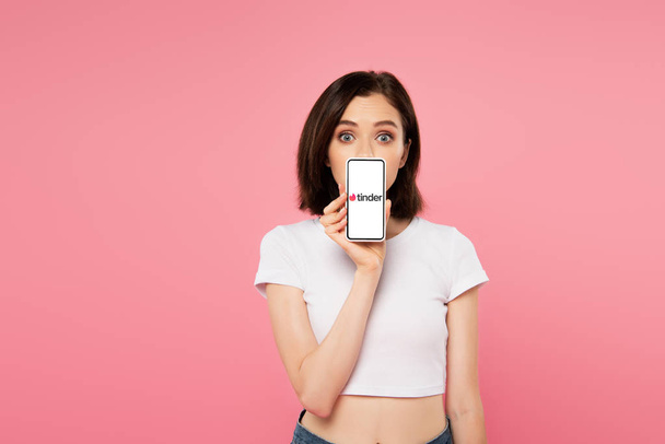 KYIV, UKRAINE - JULY 3, 2019: shocked girl holding smartphone with tinder logo isolated on pink - Fotó, kép