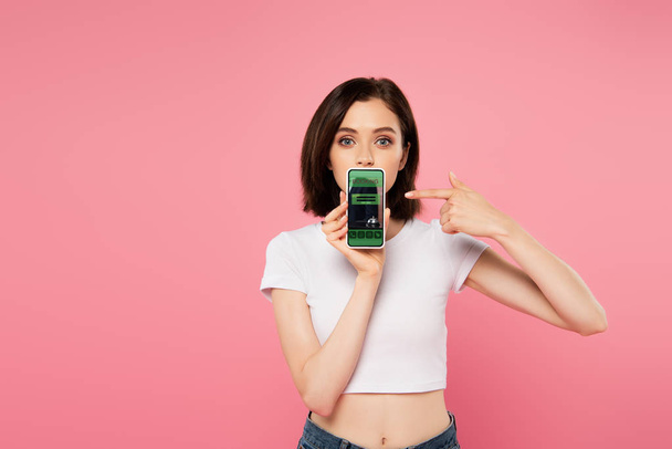 shocked girl pointing with finger at smartphone with booking app isolated on pink - Foto, immagini