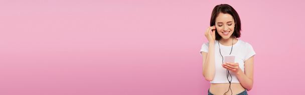 ピンクに隔離されたスマートフォンでイヤホンで音楽を聴いている笑顔の女の子のパノラマショット  - 写真・画像