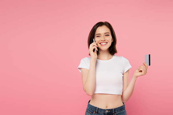 smiling girl holding credit card and talking on smartphone isolated on pink - Photo, Image