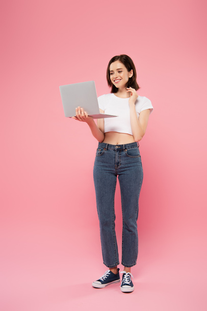full length view of smiling pretty girl holding laptop isolated on pink - Photo, image