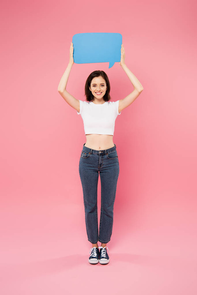 full length view of smiling pretty girl holding blank blue speech bubble isolated on pink - 写真・画像