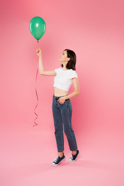 full length view of smiling pretty girl looking at green balloon isolated on pink - Foto, Imagem