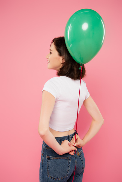 smiling pretty girl holding green balloon behind back isolated on pink - Foto, imagen