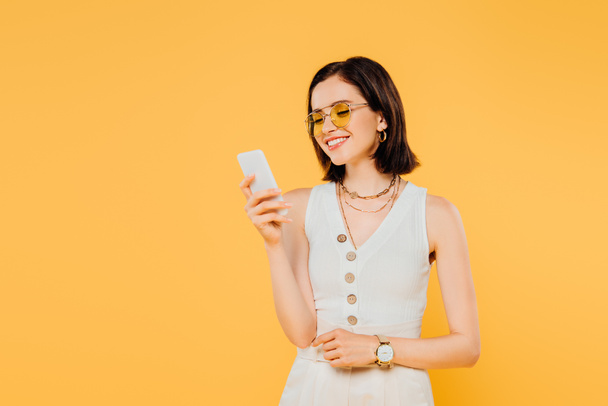 smiling elegant woman in sunglasses using smartphone isolated on yellow - Foto, Imagem