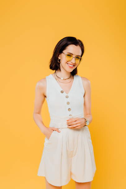 smiling stylish woman in sunglasses posing with hand in pocket isolated on yellow - Фото, зображення