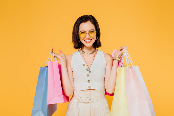 smiling woman in sunglasses holding shopping bags isolated on yellow - Photo, Image