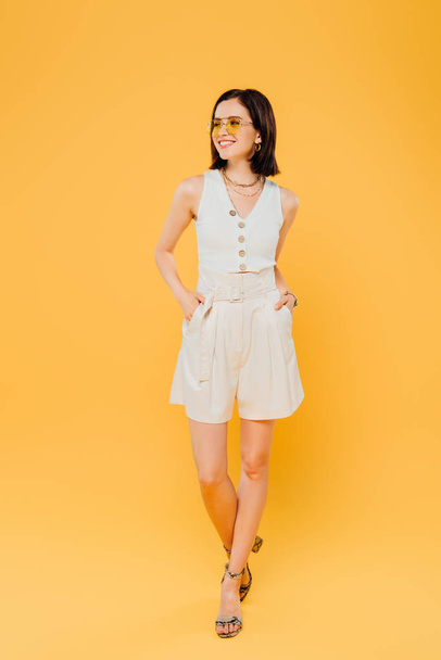 full length view of smiling fashionable girl in sunglasses with hands in pockets isolated on yellow - 写真・画像