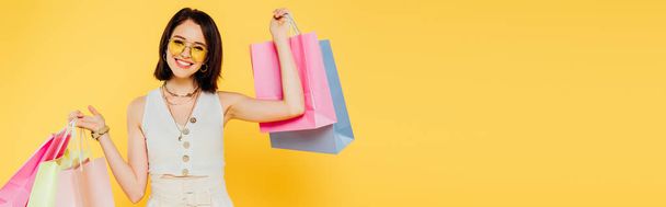 panoramic shot of happy fashionable girl in sunglasses holding shopping bags isolated on yellow - Photo, Image