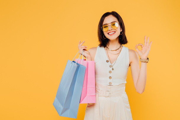 happy fashionable girl in sunglasses holding shopping bags and showing okay sign isolated on yellow - Foto, Imagem