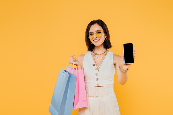 happy fashionable girl in sunglasses with shopping bags showing smartphone with blank screen isolated on yellow - Fotó, kép