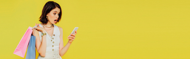 panoramatický záběr šokované dívky s nákupními sáčky, které mluví o smartphone izolovaných na žluté - Fotografie, Obrázek