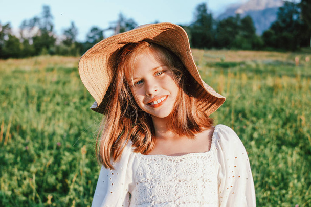 Beautiful romantic preteen girl in straw hat against the background of beautiful houses in mountain, rural scene at sunset - Photo, Image