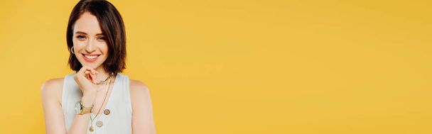 panoramatický záběr usmívající se elegantní dívky s rukou na bradu izolovanou na žluté - Fotografie, Obrázek