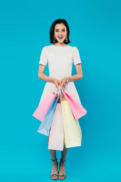 full length view of smiling elegant woman in dress with shopping bags isolated on blue - Foto, imagen
