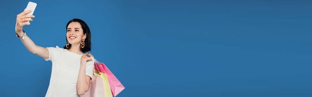 plano panorámico de mujer elegante sonriente en vestido con bolsas de compras tomando selfie en el teléfono inteligente aislado en azul
 - Foto, imagen