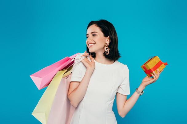 mosolygós elegáns nő ruhát bevásárló táskák gazdaság díszdobozban, és elnéz elszigetelt kék - Fotó, kép