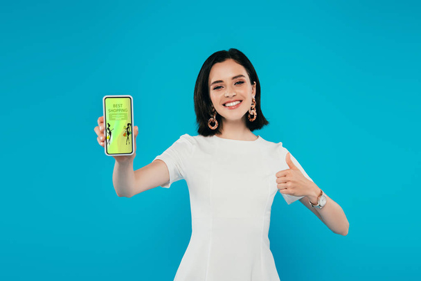 sonriente mujer elegante en vestido sosteniendo teléfono inteligente con la aplicación de compras de moda y mostrando el pulgar hacia arriba aislado en azul
 - Foto, imagen