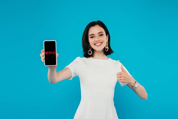 KYIV, UKRAINE - JULY 3, 2019: smiling elegant woman in dress holding smartphone with Netflix logo and showing thumb up isolated on blue - Zdjęcie, obraz