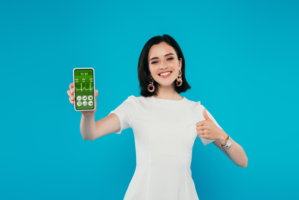 smiling elegant woman in dress holding smartphone with healthcare app and showing thumb up isolated on blue - Fotografie, Obrázek