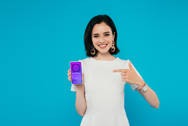 smiling elegant woman in dress pointing with finger at smartphone with online shopping app isolated on blue - Fotografie, Obrázek