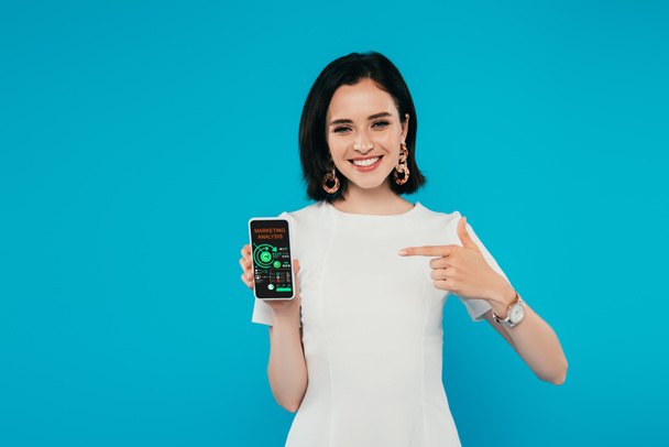 smiling elegant woman in dress pointing with finger at smartphone with marketing analysis app isolated on blue - Photo, Image