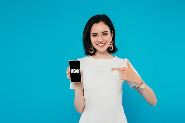 KYIV, UKRAINE - JULY 3, 2019: smiling elegant woman in dress pointing with finger at smartphone with HBO logo isolated on blue - Fotografie, Obrázek