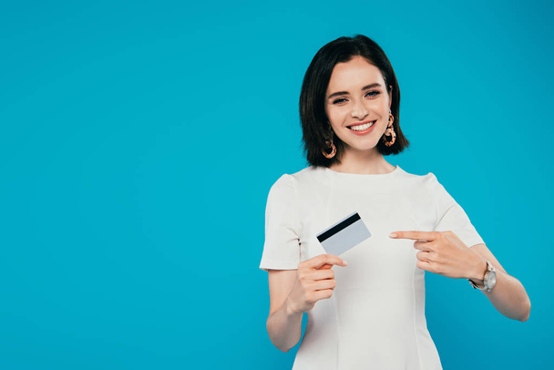 smiling elegant woman in dress pointing with finger at credit card isolated on blue - Foto, imagen