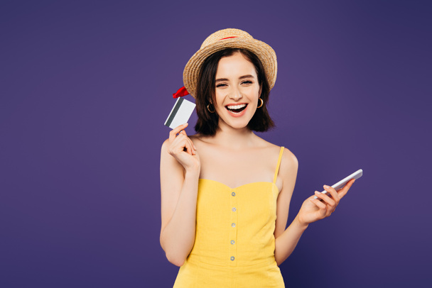 glimlachend meisje in stro hoed houden smartphone en creditcard geïsoleerd op paars - Foto, afbeelding