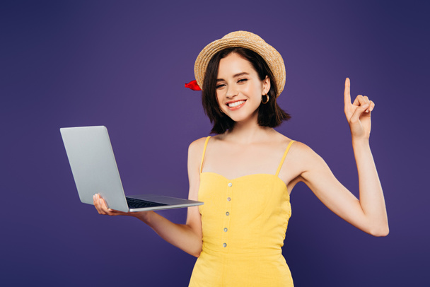 smiling girl in straw hat holding laptop and showing idea gesture isolated on purple - Zdjęcie, obraz