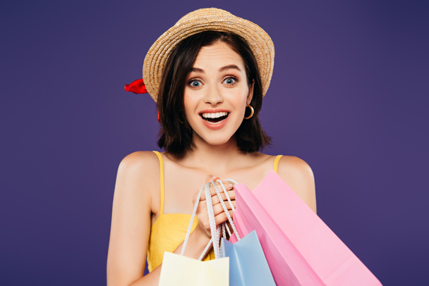 vzrušená usměvavá dívka v slaměném klobouku s nákupními pytli izolovanými na purpurovém - Fotografie, Obrázek