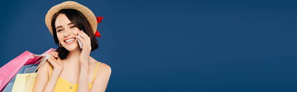 tiro panorâmico de menina sorridente em chapéu de palha com sacos de compras falando no smartphone isolado em azul
 - Foto, Imagem