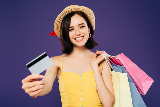 χαμογελαστή κορίτσι με ψάθινο καπέλο με σακούλες αγορών που δείχνουν πιστωτική κάρτα απομονώνονται σε μωβ - Φωτογραφία, εικόνα