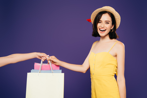 chica sonriente en sombrero de paja tomando bolsas de compras aisladas en púrpura
 - Foto, Imagen