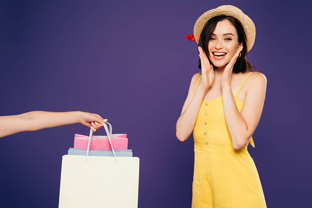 smiling girl in straw hat rejoicing near woman holding shopping bags isolated on purple - Foto, Imagem