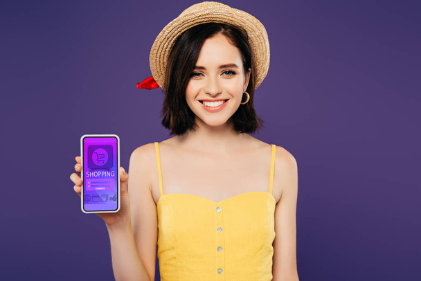 chica sonriente en sombrero de paja celebración de teléfono inteligente con aplicación de compras aislado en púrpura
 - Foto, imagen