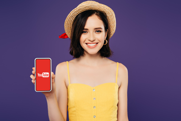 KYIV, UKRAINE - JULY 3, 2019: smiling girl in straw hat holding smartphone with youtube app isolated on purple - Photo, image