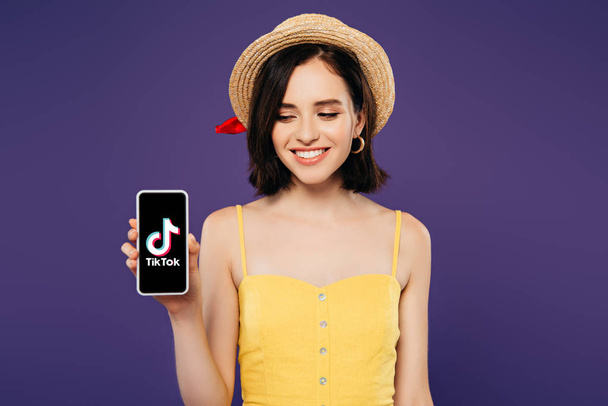 KYIV, UKRAINE - JULY 3, 2019: smiling girl in straw hat holding smartphone with tiktok app isolated on purple - Photo, Image