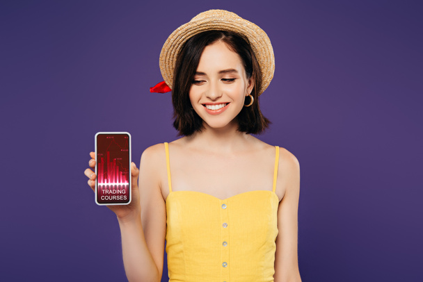 усміхнена дівчина в солом'яному капелюсі тримає смартфон із додатком торгових курсів ізольовано на фіолетовому
 - Фото, зображення
