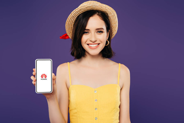 KYIV, UKRAINE - JULY 3, 2019: smiling girl in straw hat holding smartphone with huawe logo isolated on purple - Fotó, kép