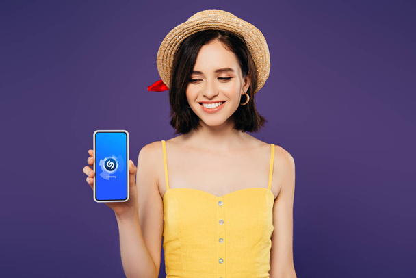 KYIV, UKRAINE - JULY 3, 2019: smiling girl in straw hat holding smartphone with shazam app isolated on purple - Photo, Image