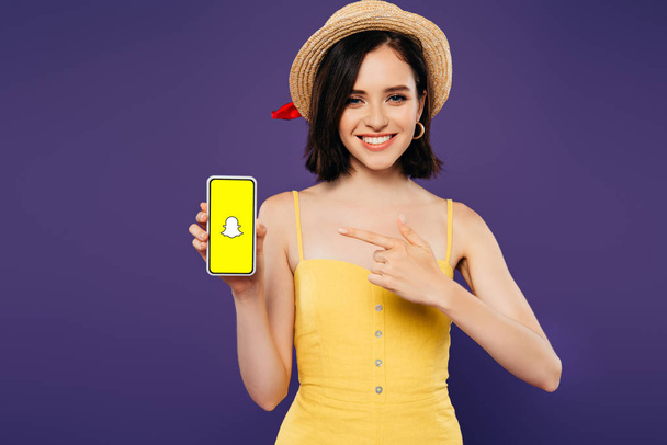 KYIV, UKRAINE - JULY 3, 2019: smiling girl in straw hat pointing with finger at smartphone with snapchat app isolated on purple - Foto, Imagem