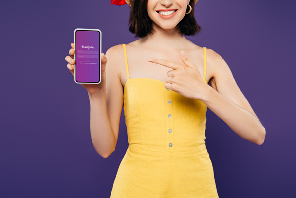 KYIV, UKRAINE - JULY 3, 2019: cropped view of girl in straw hat pointing with finger at smartphone with instagram app isolated on purple - Foto, Bild