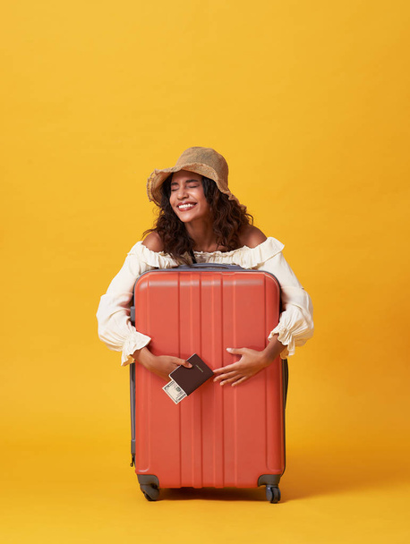 Allegro giovane donna africana vestita in abiti estivi in possesso di passaporto con i soldi mentre abbraccio con valigia isolato su sfondo giallo
. - Foto, immagini