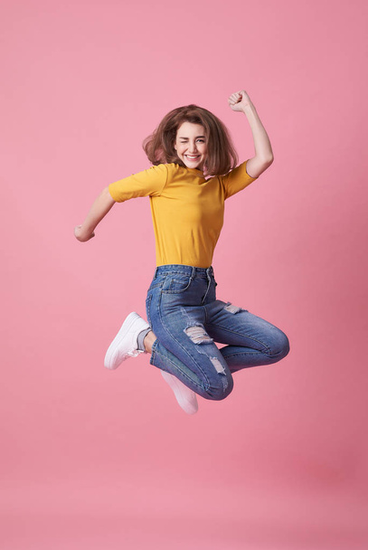 Portrait of a joyful young woman in yellow shirt jumping and celebrating over pink background. - Fotografie, Obrázek