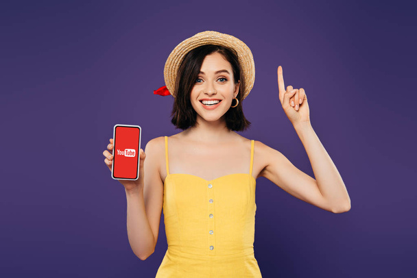 КИЕВ, Украина - 3 июля 2019 года: взволнованная девушка в соломенной шляпе, показывающая жест идеи и держащая смартфон с приложением youtube изолированным на фиолетовый
 - Фото, изображение