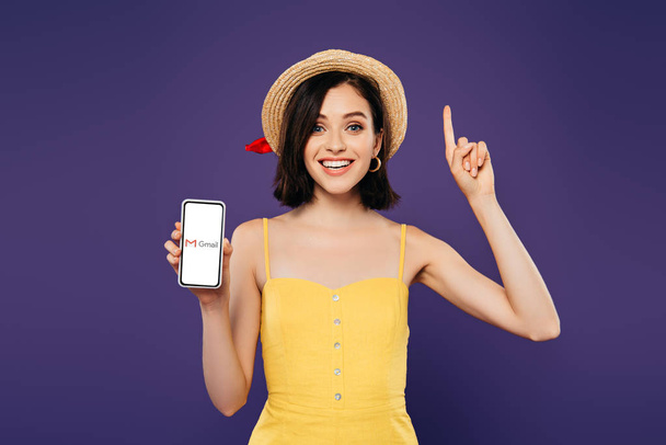KYIV, UKRAINE - JULY 3, 2019: excited girl in straw hat showing idea gesture and holding smartphone with gmail app isolated on purple - Фото, изображение