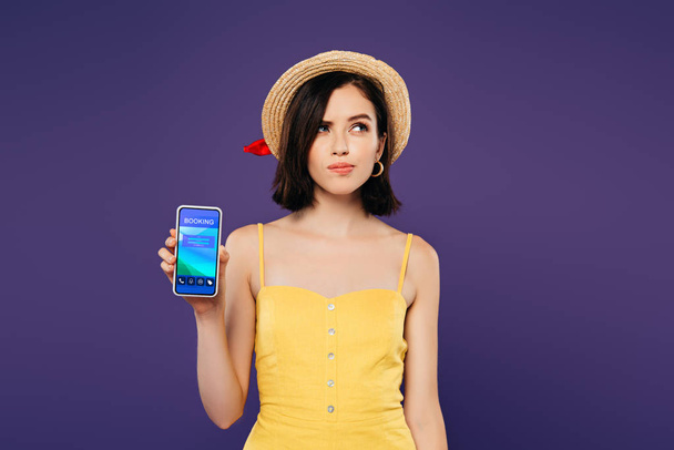 verträumtes hübsches Mädchen mit Strohhut hält Smartphone mit Buchungs-App isoliert auf lila  - Foto, Bild