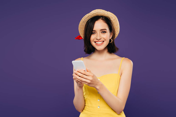 sonriente chica bonita en sombrero de paja usando teléfono inteligente aislado en púrpura
  - Foto, imagen