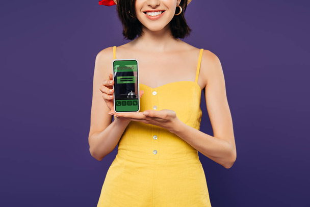 cropped view of smiling girl holding smartphone with booking app isolated on purple  - Fotografie, Obrázek